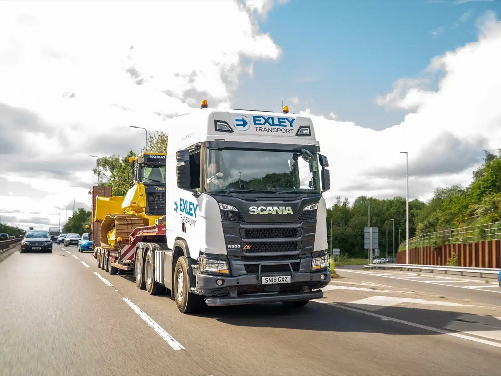 Liveried low loader lorry carrying plant on the road